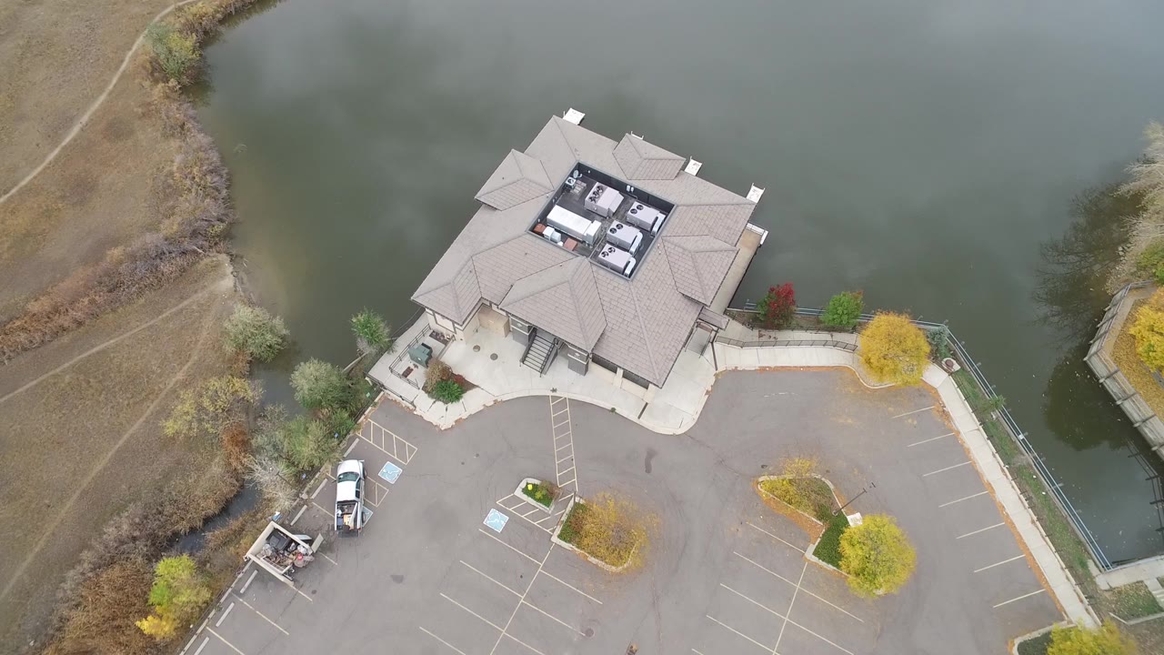 Aerial frame in Hidden Lake, Colorado showing the building roof closer to center, the water curving behind it, empty parking stalls, several parked vehicles, and small landscaped trees.