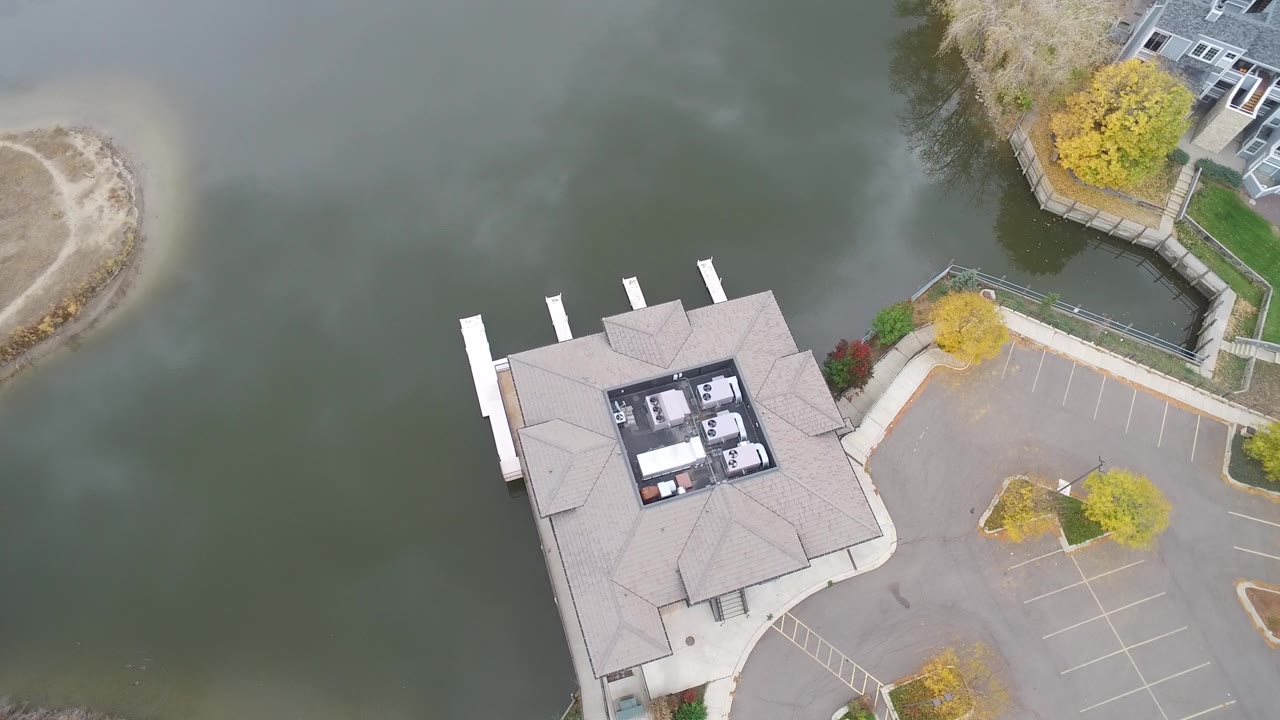 Overhead frame from Hidden Lake, Colorado showing green lake water beside a roofed lakeside business, white dock slips, a mostly empty parking lot, shoreline paths, and no visible people.