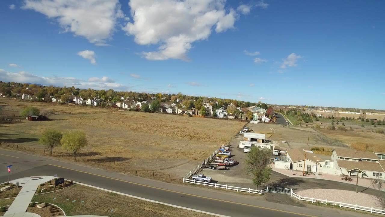 Aerial view of road, fenced buildings, parked cars, open field, and houses under blue sky.