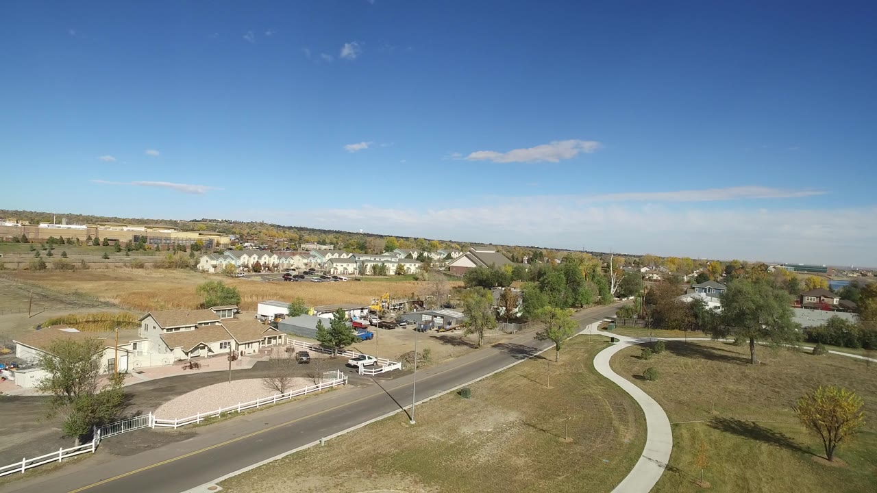 Opening aerial frame over Hidden Lake, Colorado, showing a two-lane road, white fence, curved path, open grass, trees, distant homes, and no visible people.