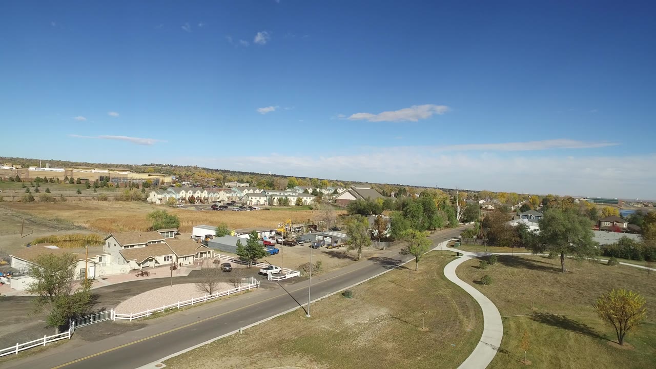 Wide aerial view of parkland, a roadside fence, paths, trees, and suburban buildings.