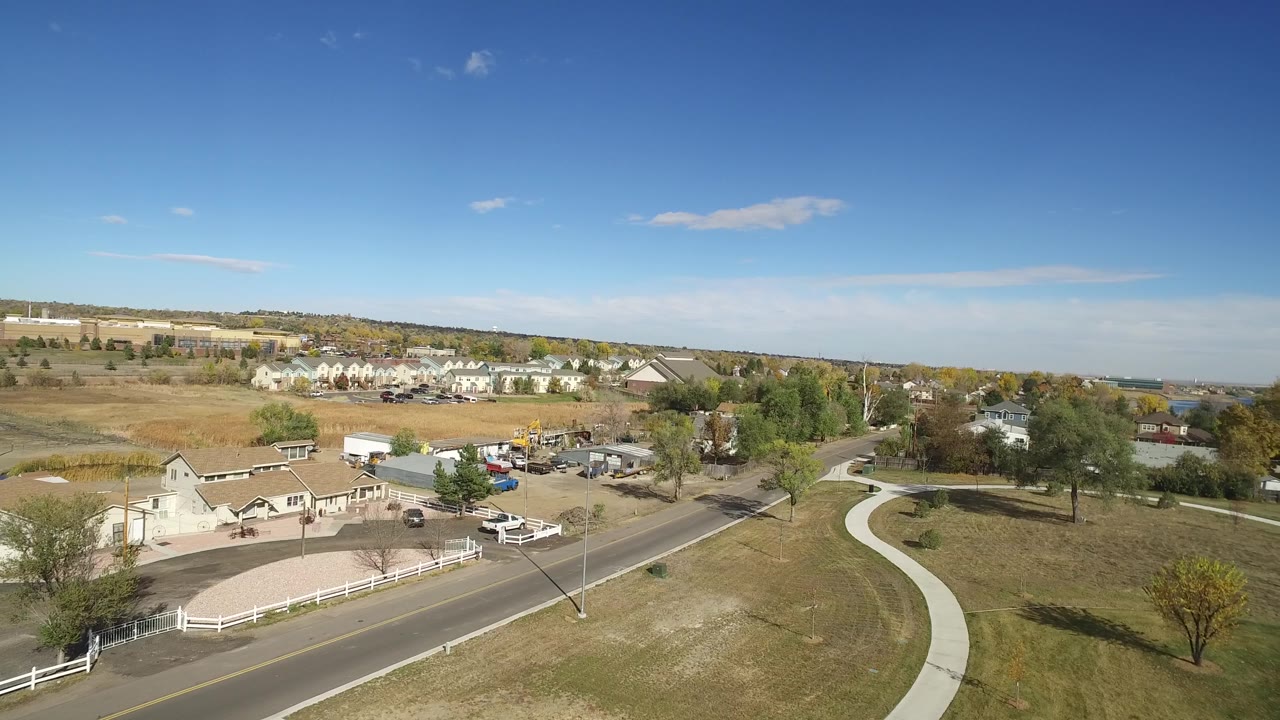 Hidden Lake, Colorado aerial frame with a curving walkway at right, open lawn, roadside intersection area, scattered trees, distant houses and buildings, and no visible people.