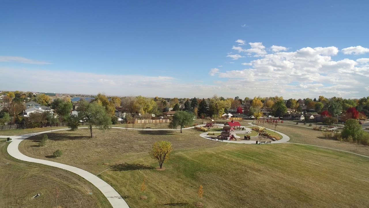 Aerial view of a Hidden Lake, Colorado park with a large grass field in front, curving concrete paths, a red-roofed play area, autumn trees, distant homes, and small people visible near the park center.