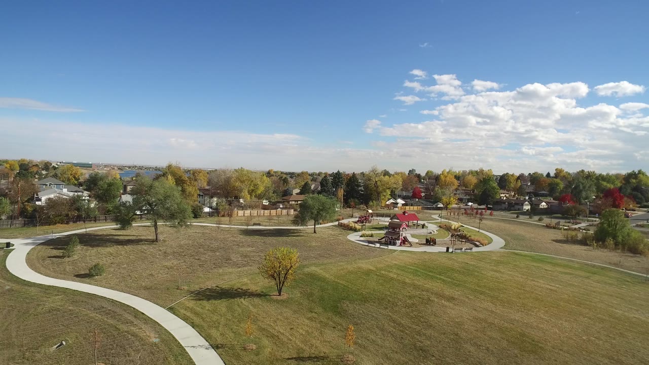 A wide aerial frame in Hidden Lake, Colorado shows the walking path sweeping across the foreground grass toward a red-roofed park feature, tree-lined homes, and tiny people in the distance.