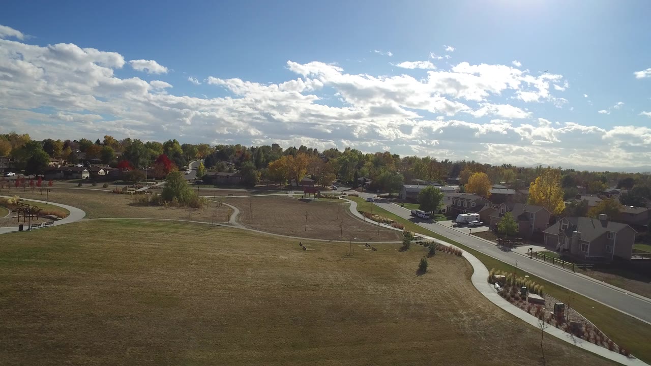 Elevated view over grass, paths, roads, homes, trees, and bright clouds.