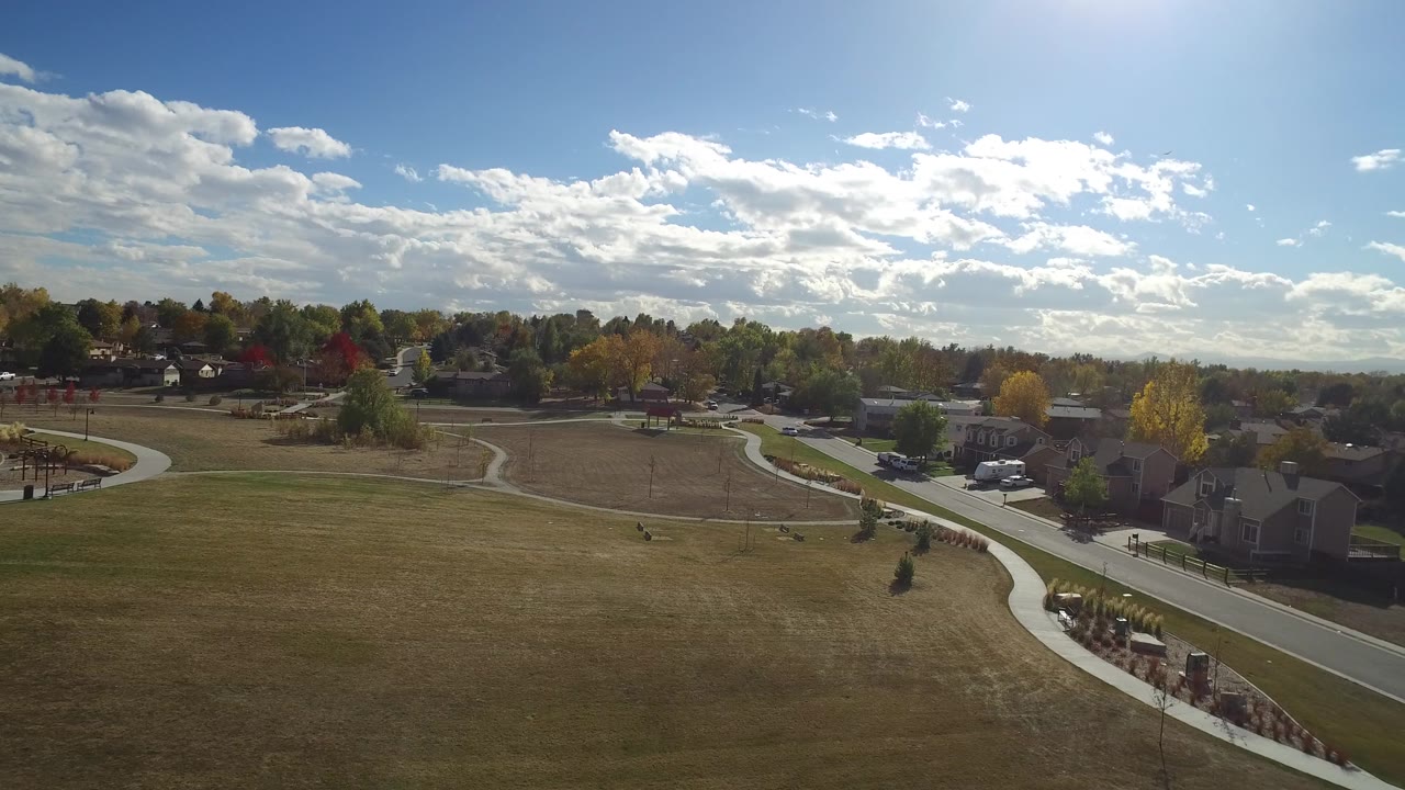At 0:01, the Hidden Lake aerial view centers on a curving sidewalk through open grass with houses, parked vehicles, autumn trees, and bright clouds beyond.