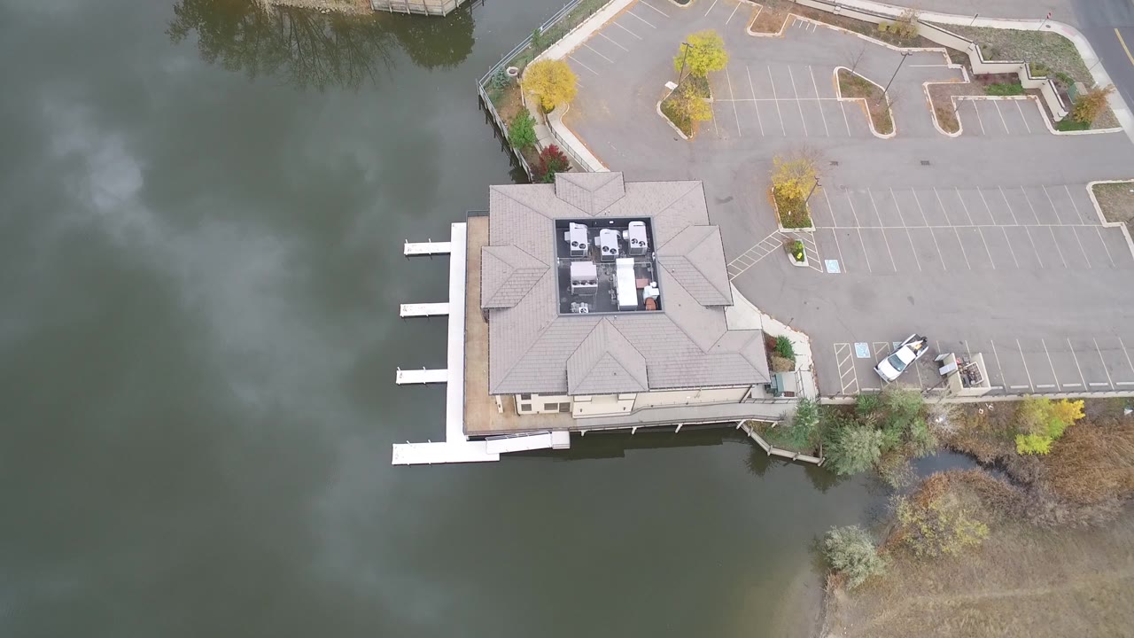 Downward drone view at Hidden Lake, Colorado with cloudy reflections on the water, the lakeside building near the lower center, dock slips along the shore, a mostly empty parking lot, and no visible people.
