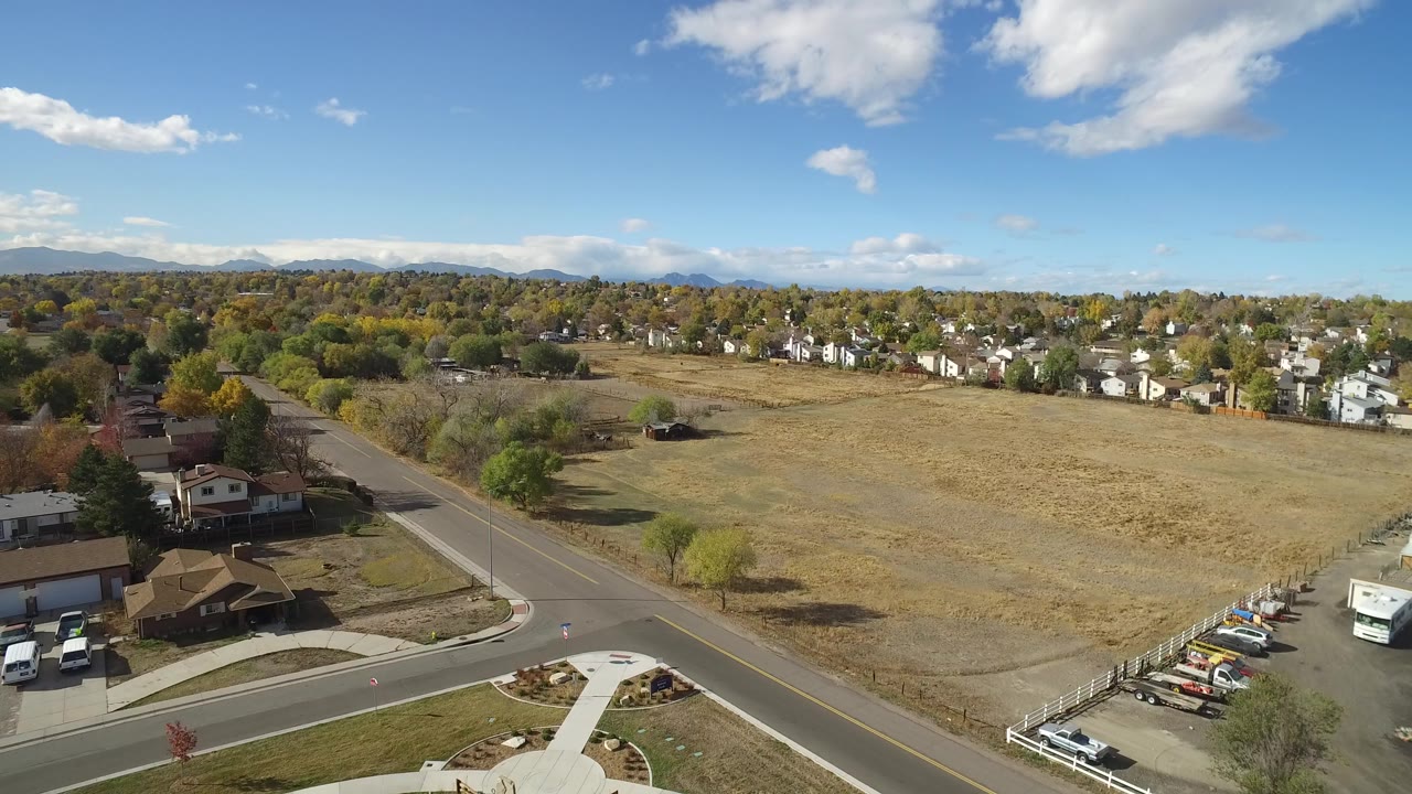 At 0:00, an elevated view in Hidden Lake, Colorado shows a road curving past a dry field, nearby homes, autumn trees, distant mountains, and no visible people.