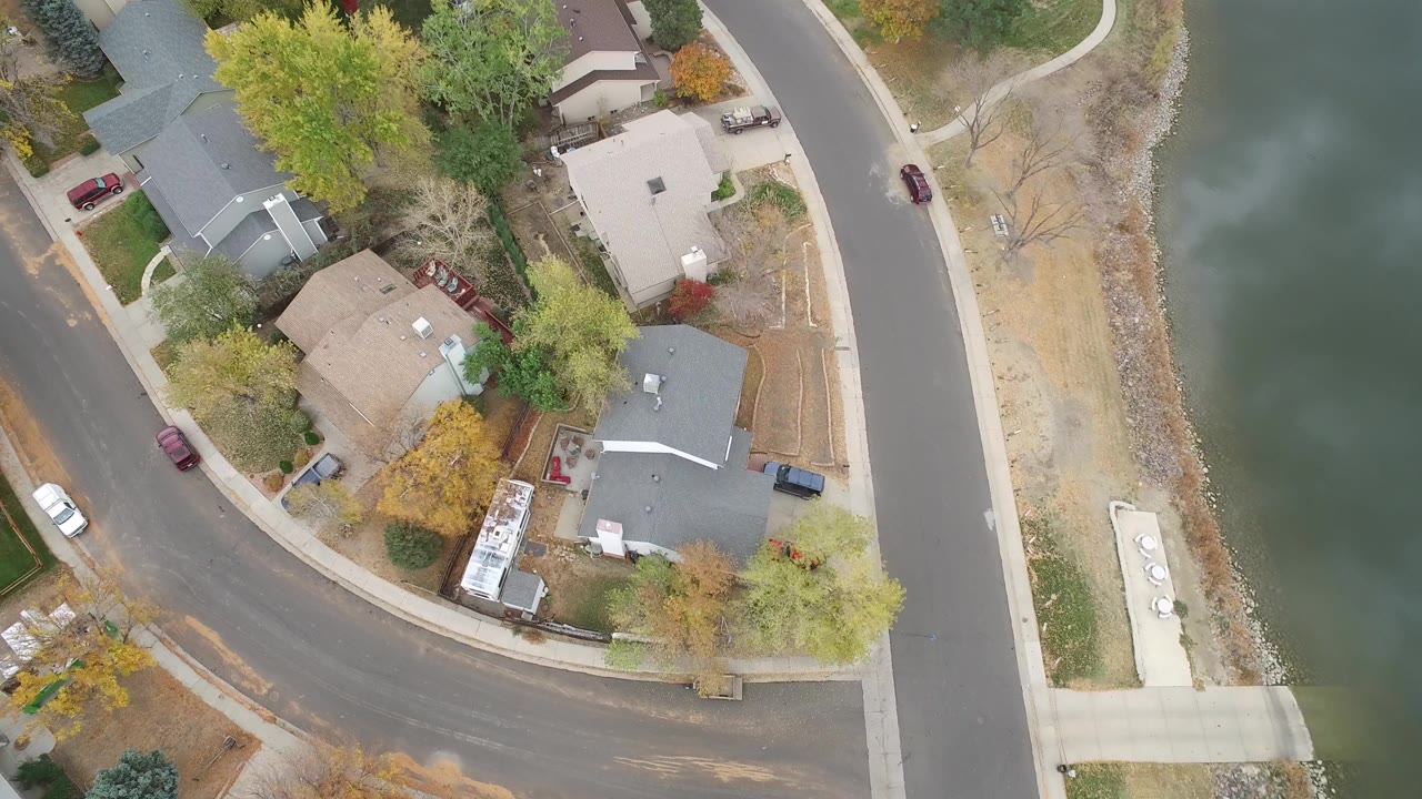 Overhead frame from Hidden Lake, Colorado showing detached homes along curved roads beside a strip of shoreline and green lake water, with no visible people.
