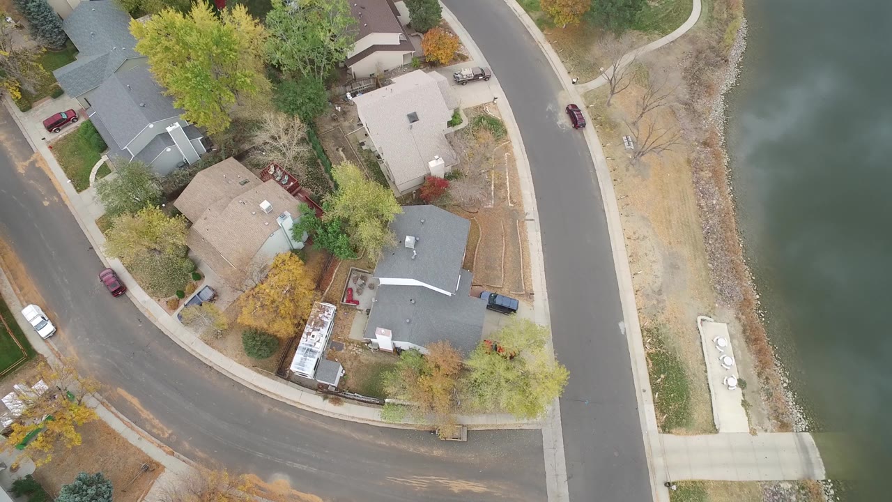 A high overhead view in Hidden Lake, Colorado centers several homes and tree cover while the lakeside road and water remain along the right side, with no visible people.