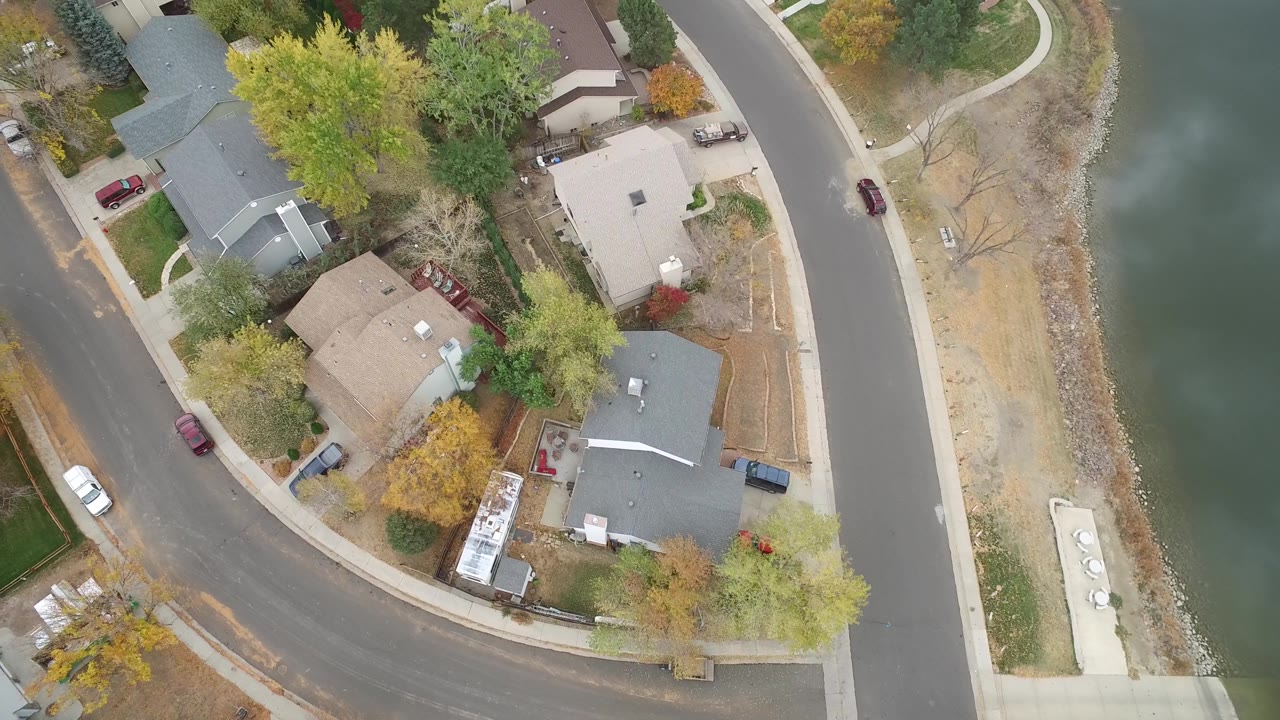 Top-down neighborhood view with houses, roads, trees, cars, and lake water.