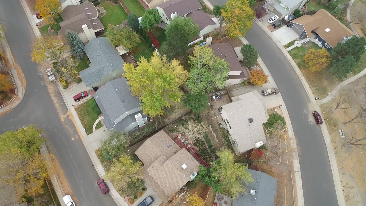 Overhead view of suburban houses, curved streets, trees, and parked cars.