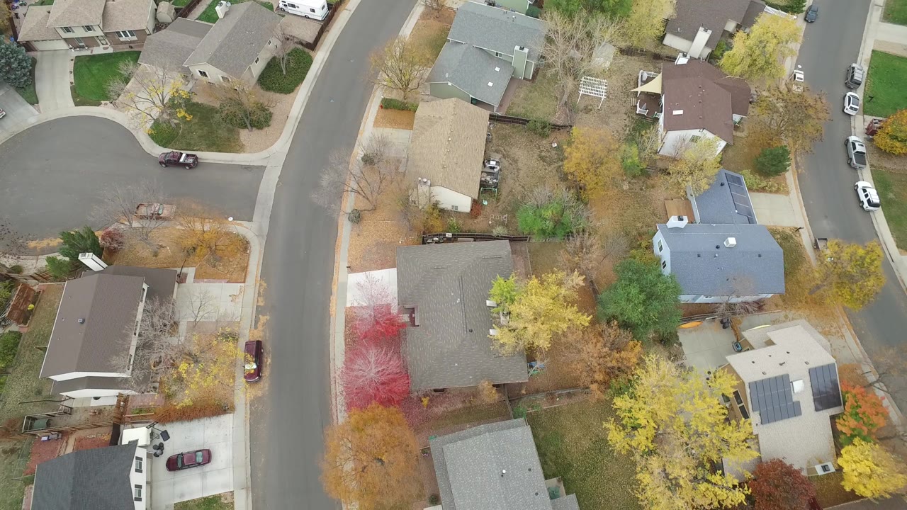 Overhead view of a curved residential street with houses, yards, trees, and parked cars.