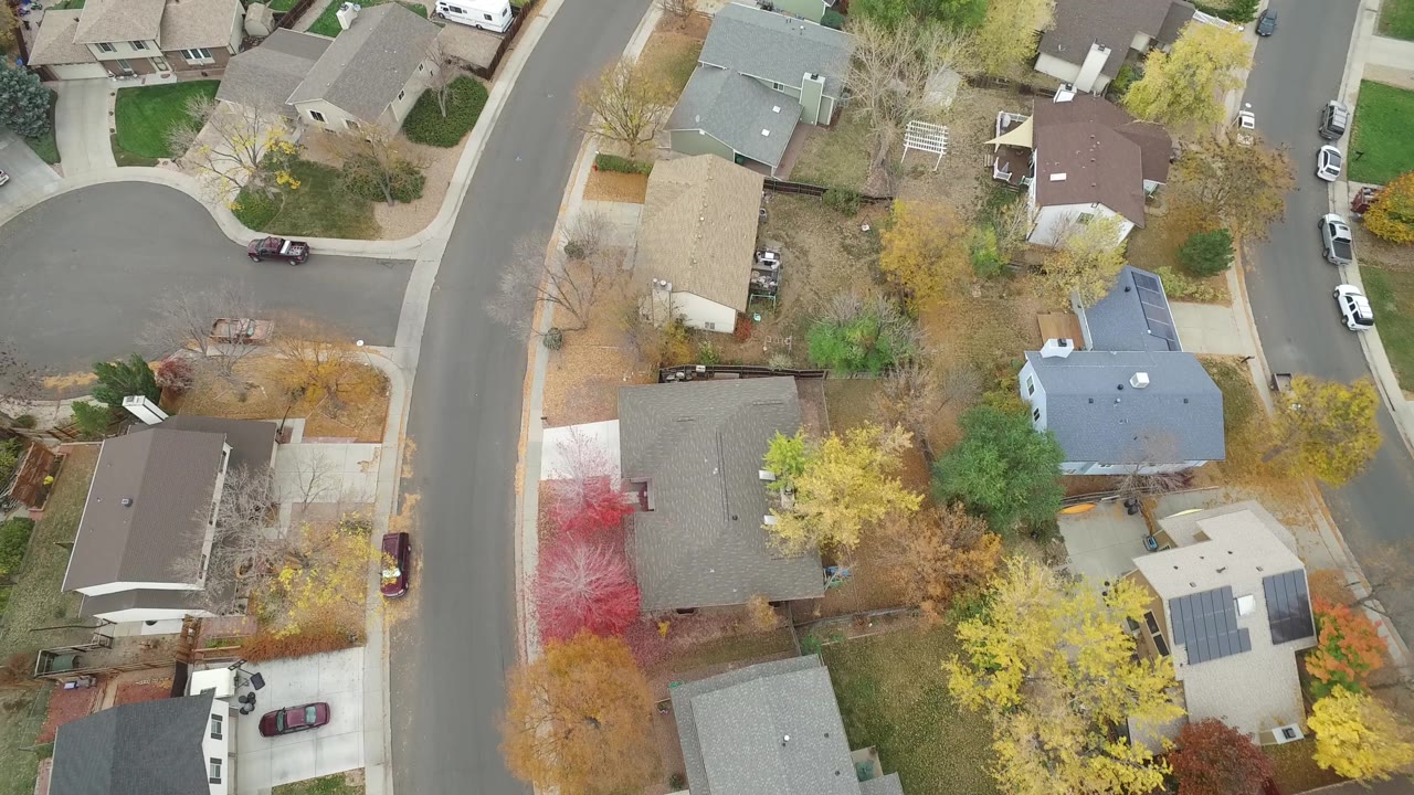 Aerial frame from Hidden Lake, Colorado with a cul-de-sac edge on the left, residential rooftops, mixed yellow and bare trees, parked cars, and no visible people.