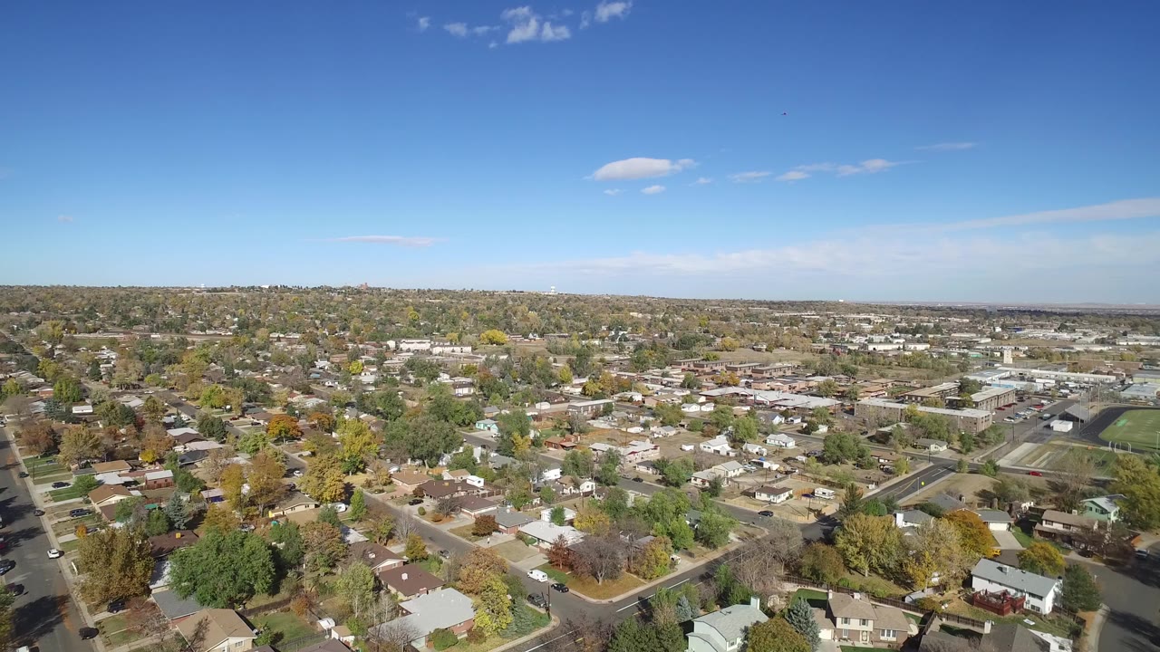 Aerial view over suburban houses and tree-lined streets under blue sky.