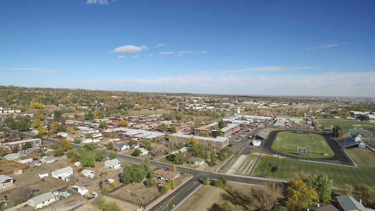 Aerial view of houses, roads, open lots, and an athletic field under blue sky.