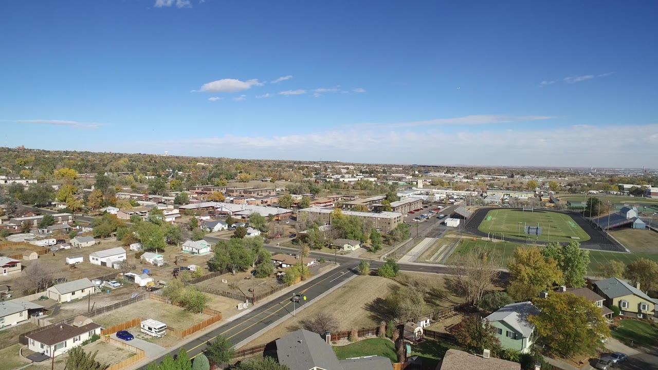 Aerial view over suburban homes, streets, trees, and a sports field under blue sky.