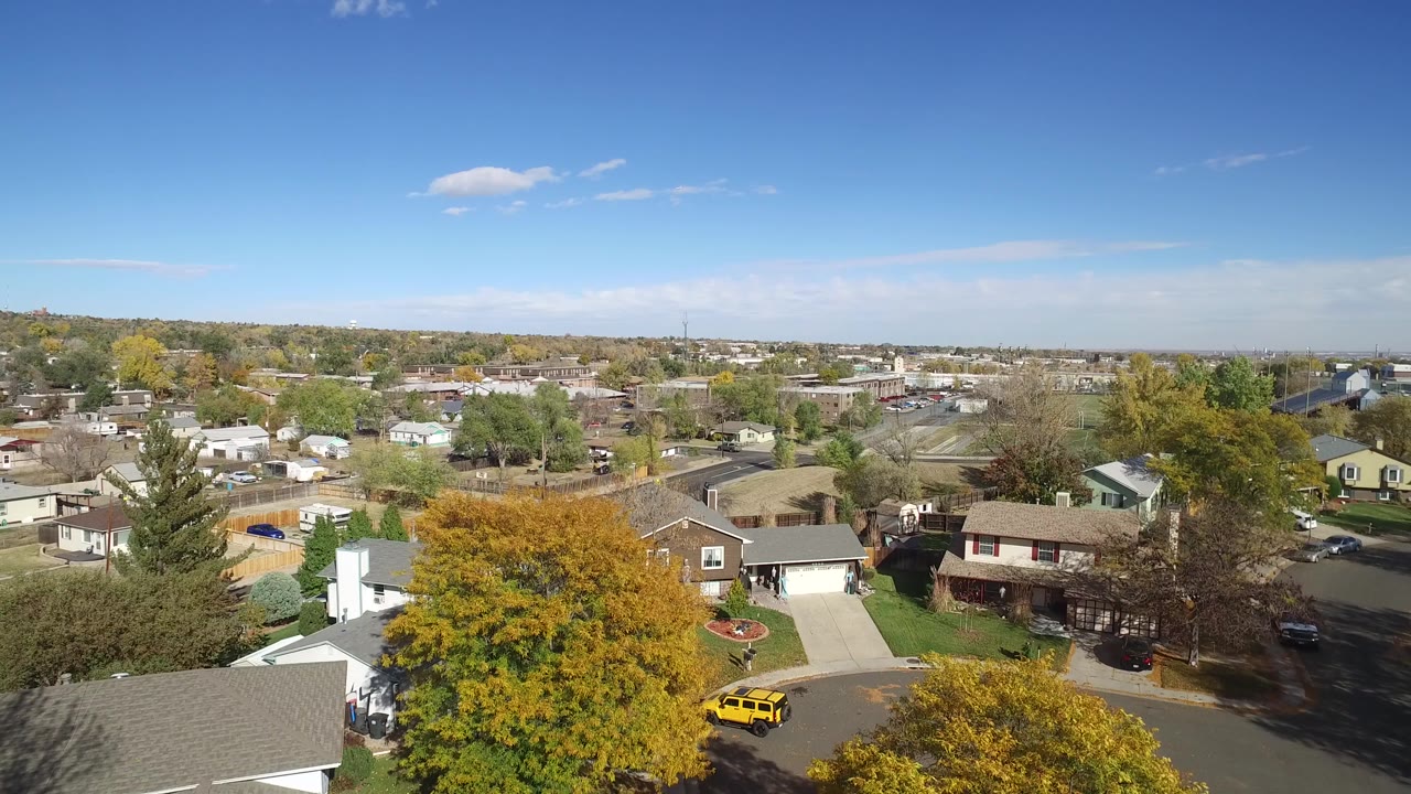 Aerial view over suburban homes, trees, streets, and blue sky.