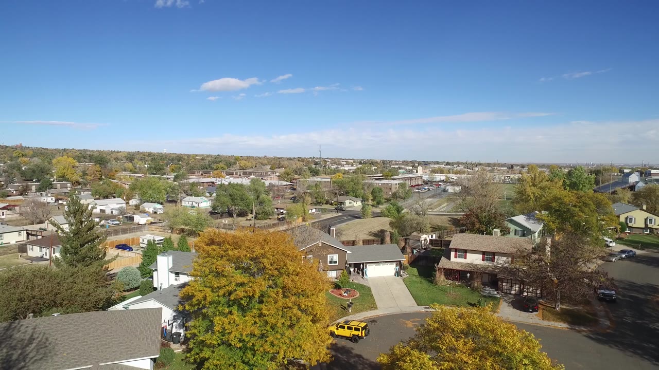 High aerial frame in Hidden Lake, Colorado showing a yellow vehicle near the street, detached homes, driveways, mature trees, open sky, and no visible people.