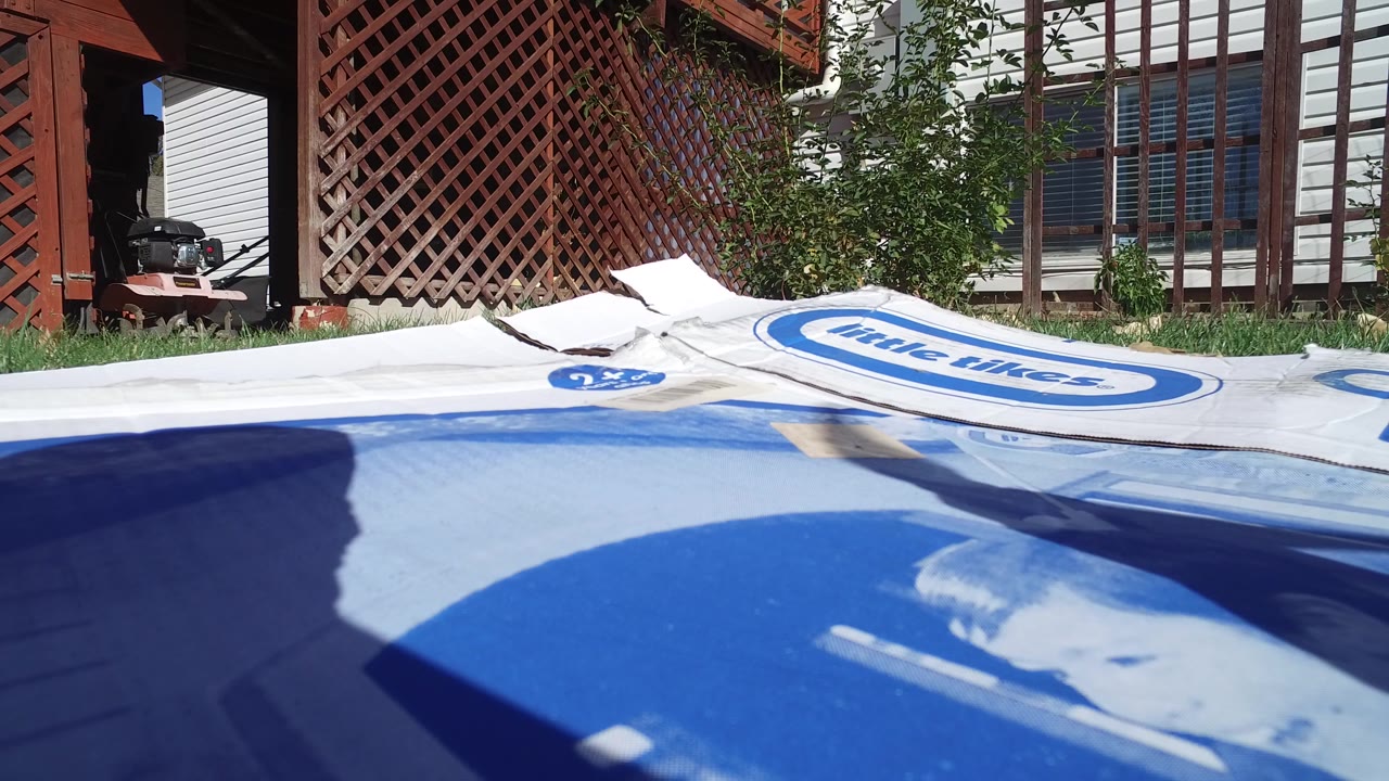 Low close view in Hidden Lake, Colorado of a blue-and-white plastic box on grass with a propeller shadow, shrubs, trellis, house siding, and no visible people.
