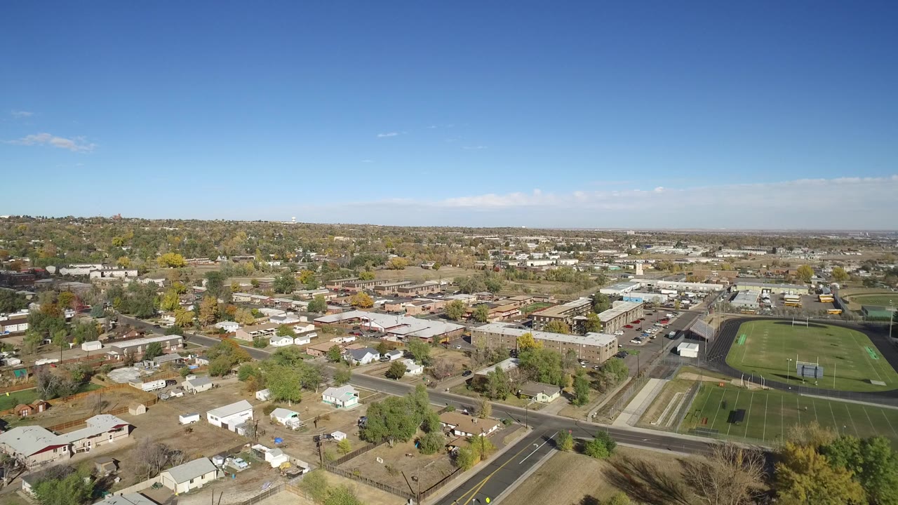 Aerial view of suburban roads, homes, low buildings, and a green athletic field under blue sky.