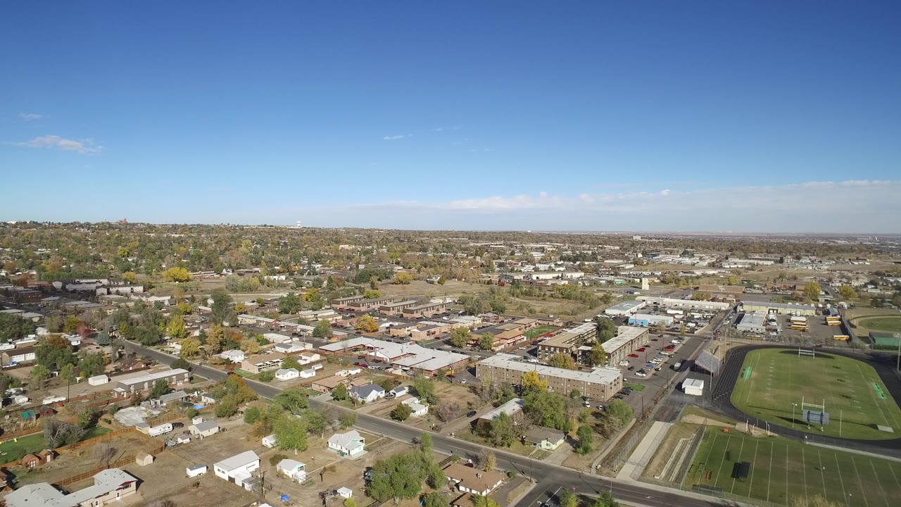 Aerial view over homes, roads, sports field, and low buildings under blue sky.