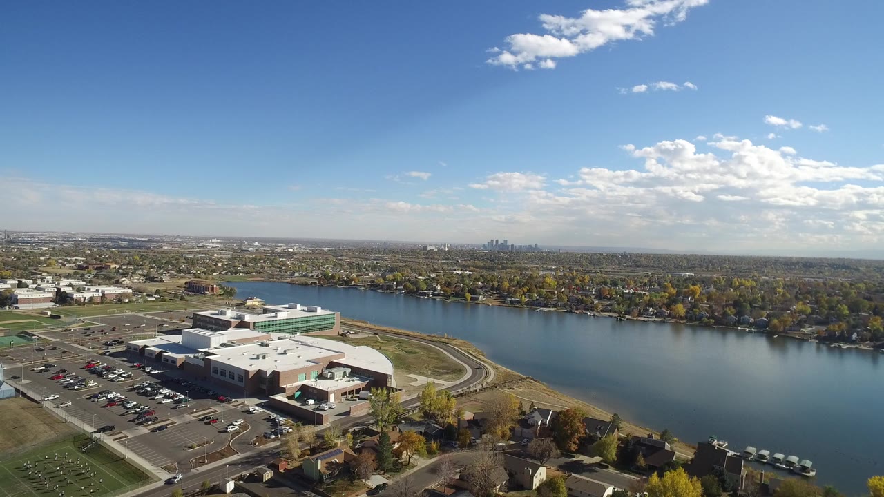 Aerial frame over Hidden Lake, Colorado showing a large lakeside building, parking lot, blue water, tree-lined homes, distant skyline, and tiny people not individually discernible.