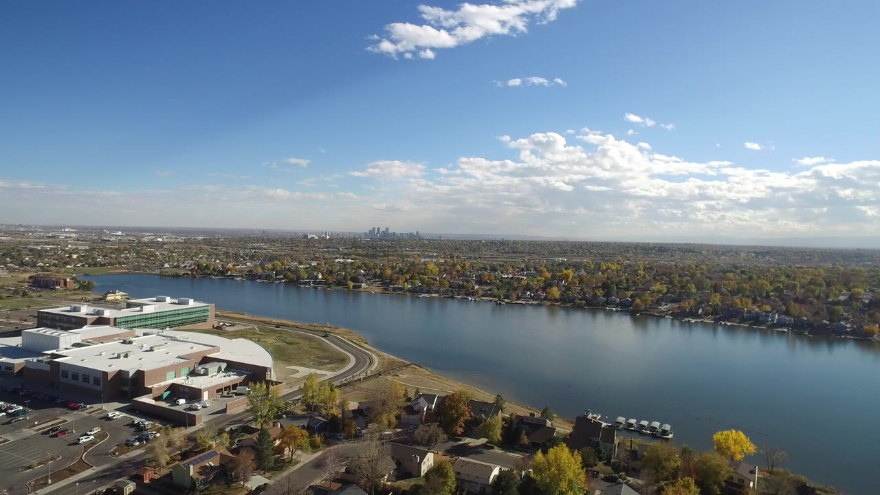 Aerial view in Hidden Lake, Colorado showing low commercial buildings in the foreground, broad blue water, tree-lined shoreline, distant skyline, scattered clouds, and no visible people.