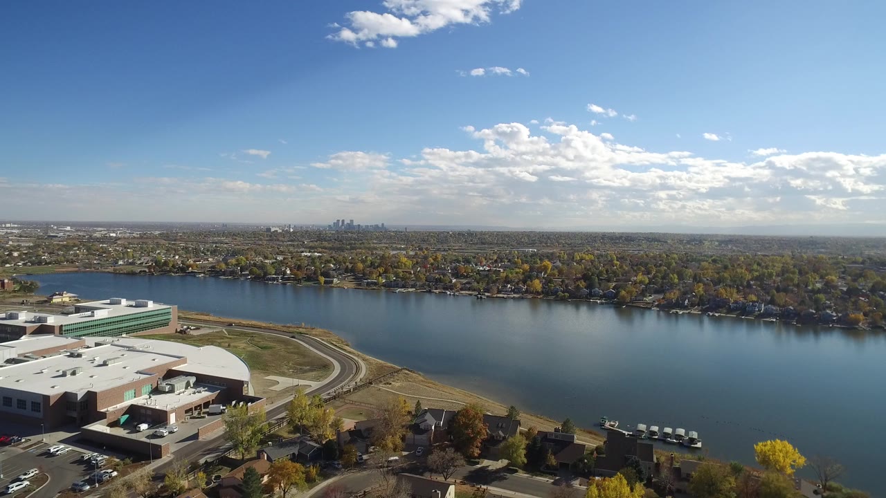 Wide aerial view over a lake, shoreline homes, roads, and a large foreground building.