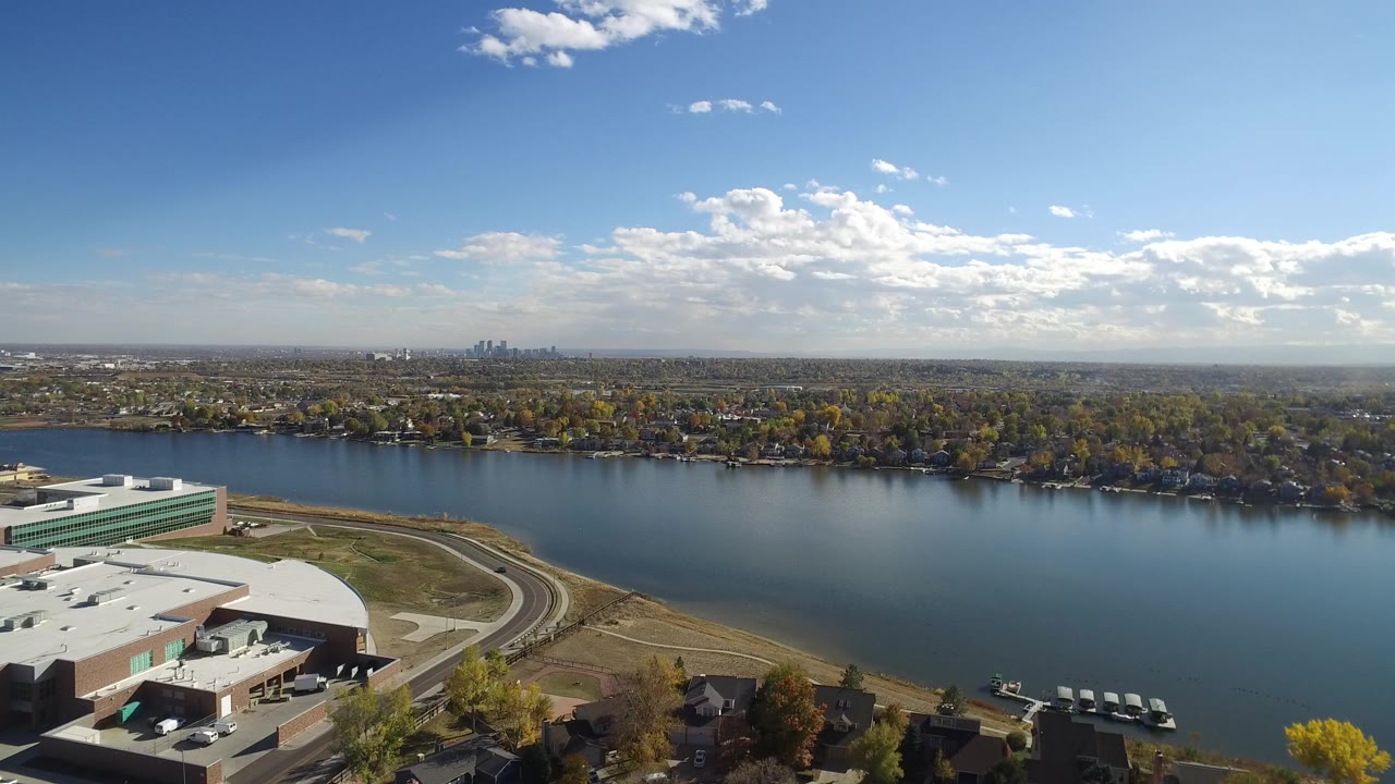 Opening aerial frame over Hidden Lake, Colorado with a white-roofed building at lower left, blue water across the center, homes and trees beyond, a distant skyline, and no visible people.