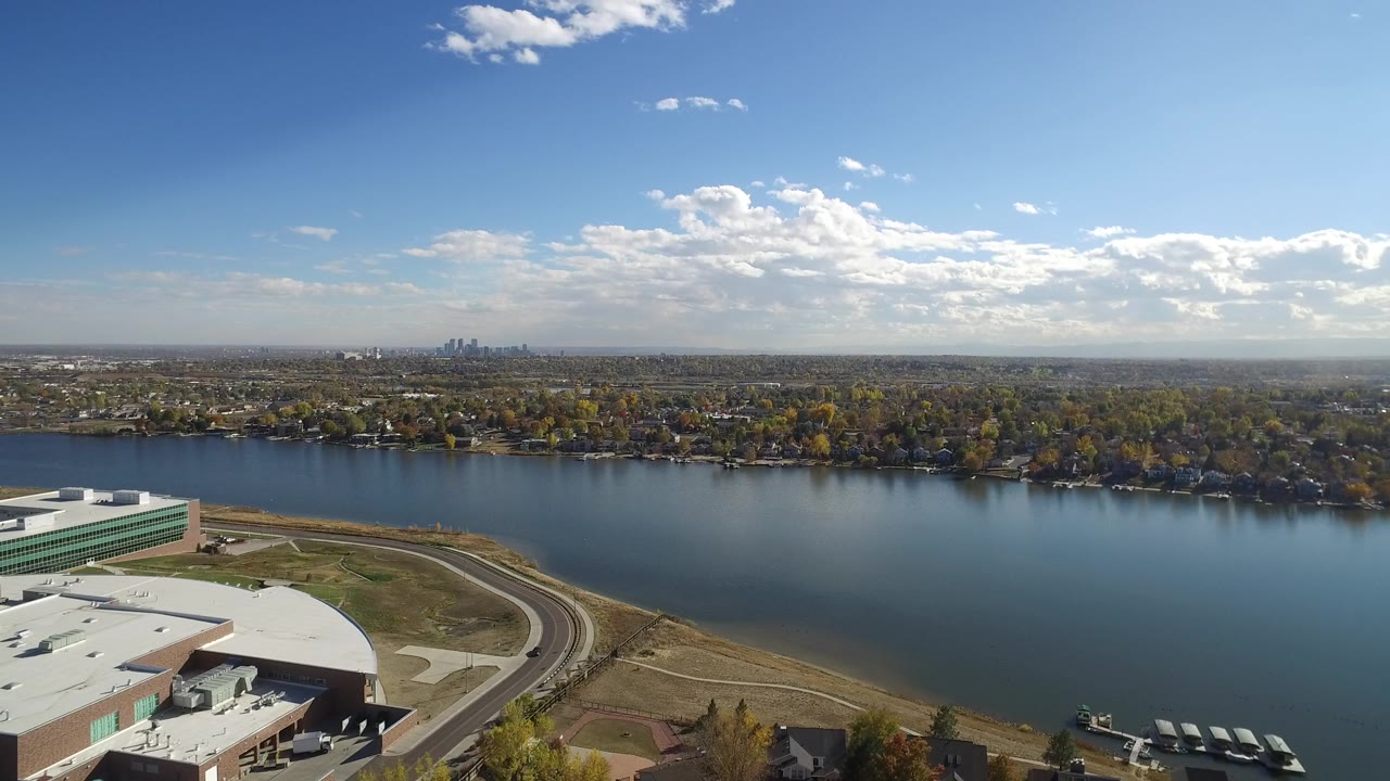 Second aerial frame over Hidden Lake, Colorado, showing the road and large building roof lower in frame, smooth blue water through the middle, autumn-colored trees and homes on the far shore, and no visible people.