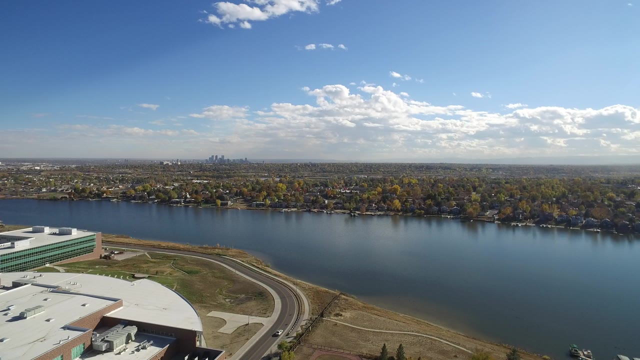 Aerial view over a white-roofed building, curved road, blue water, trees, and distant skyline.