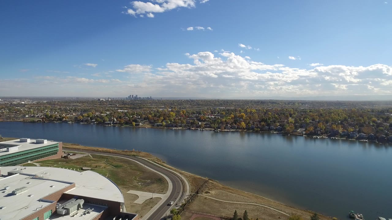 One second into the Hidden Lake, Colorado clip, the elevated view holds on the building complex, shoreline road, wide water surface, autumn-colored trees, far homes, distant skyline, and no visible people.