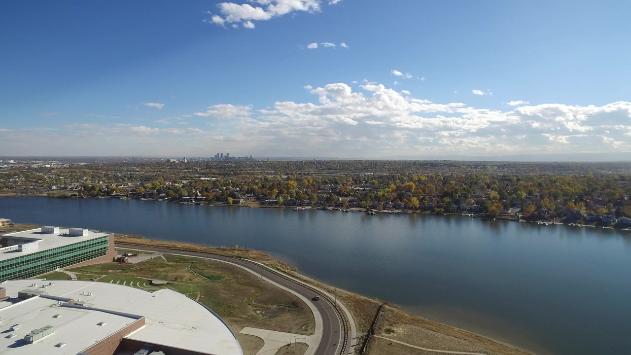 Aerial view over blue water with buildings, road, trees, and distant skyline.