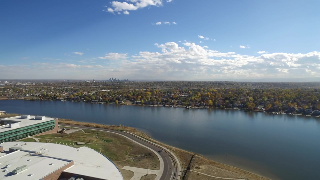Hidden Lake, Colorado aerial frame with calm blue water, foreground commercial-looking rooftops, a curving road, wooded residential shoreline, distant skyline, and no visible people.