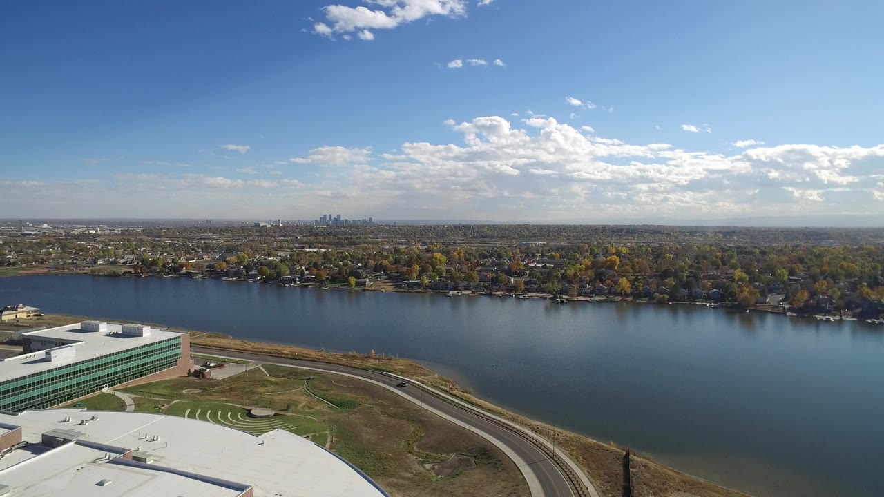 Aerial view over a lake, treed shoreline, foreground rooftops, and distant skyline under clouds.