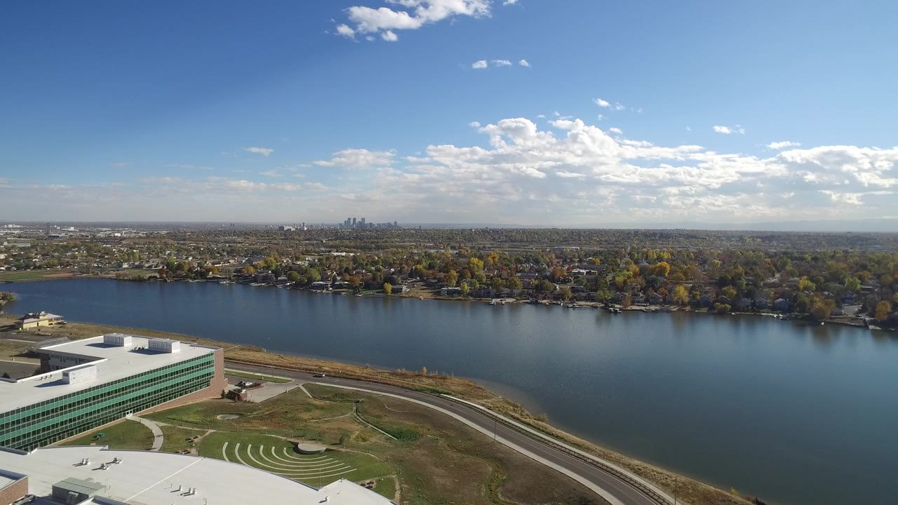 Opening aerial frame over Hidden Lake, Colorado, with a green-glass building at lower left, blue water across the center, a tree-lined shore beyond, a distant skyline on the horizon, and no visible people.