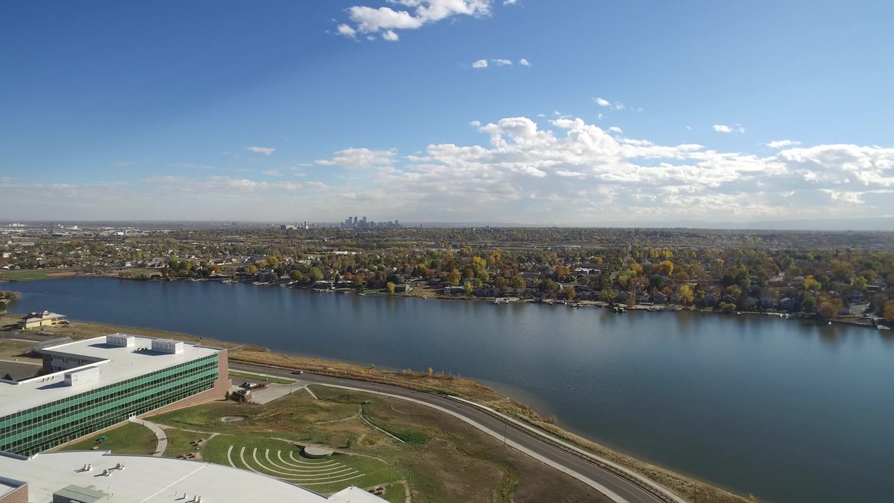 Aerial lakefront scene with building corner, shoreline road, trees, and distant city skyline.