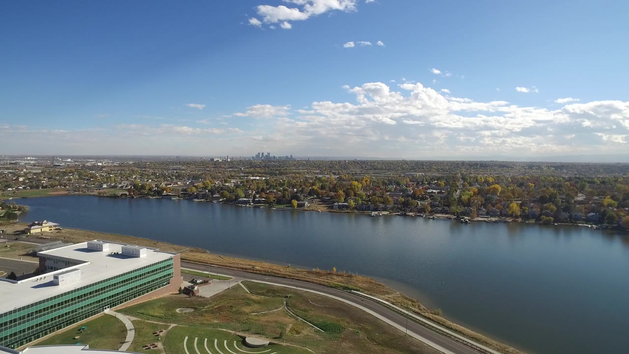 Aerial view of a blue lake, shoreline homes, curving road, and white building under a bright sky.