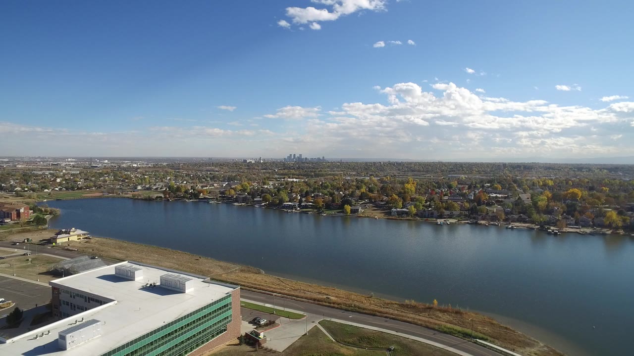 Opening aerial frame of Hidden Lake, Colorado, with a white-roofed building at lower left, blue lake across the center, residential streets beyond, distant skyline, and no visible people.