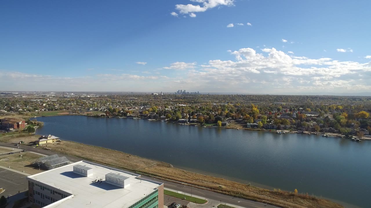 Aerial frame over Hidden Lake, Colorado with a white rooftop at lower left, blue lake water across the center, homes and trees along the far shore, distant skyline on the horizon, and no visible people.