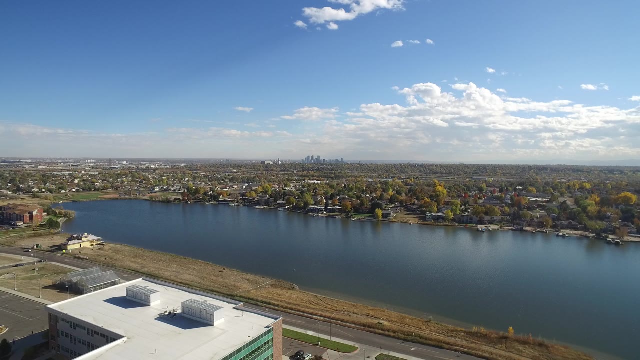 Hidden Lake, Colorado appears from above with calm blue water in the foreground, a line of shoreline trees and houses beyond it, a distant urban skyline left of center, and no visible people.
