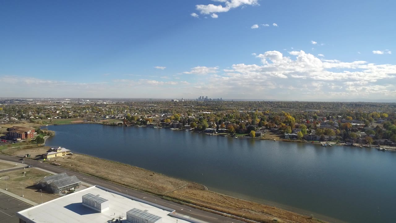 Opening aerial frame over Hidden Lake, Colorado, showing blue water across the center, a road and dry grass in front, homes and trees beyond the shore, distant skyline, and no visible people.