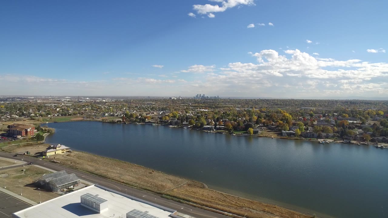 Aerial lake scene with industrial rooftop corner, road, homes, trees, and skyline under clouds.