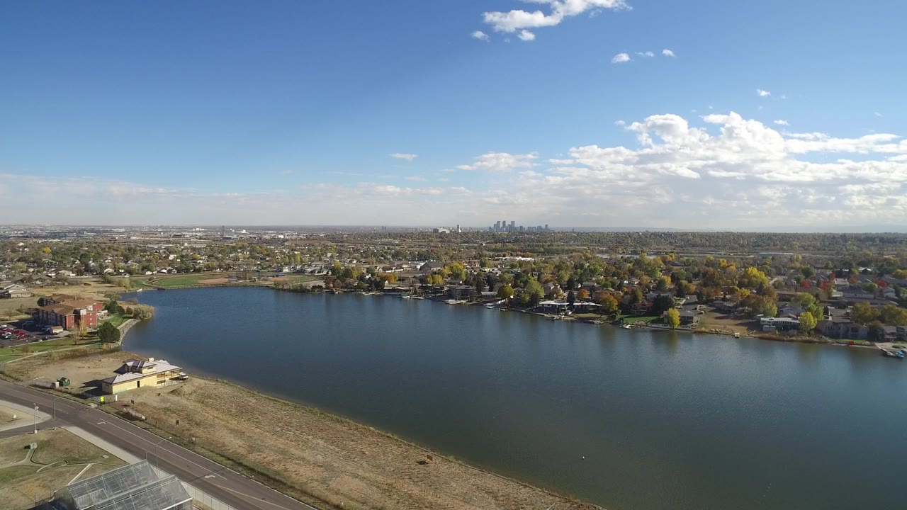Wide aerial view of a lake with shoreline roads, homes, trees, and distant skyline.