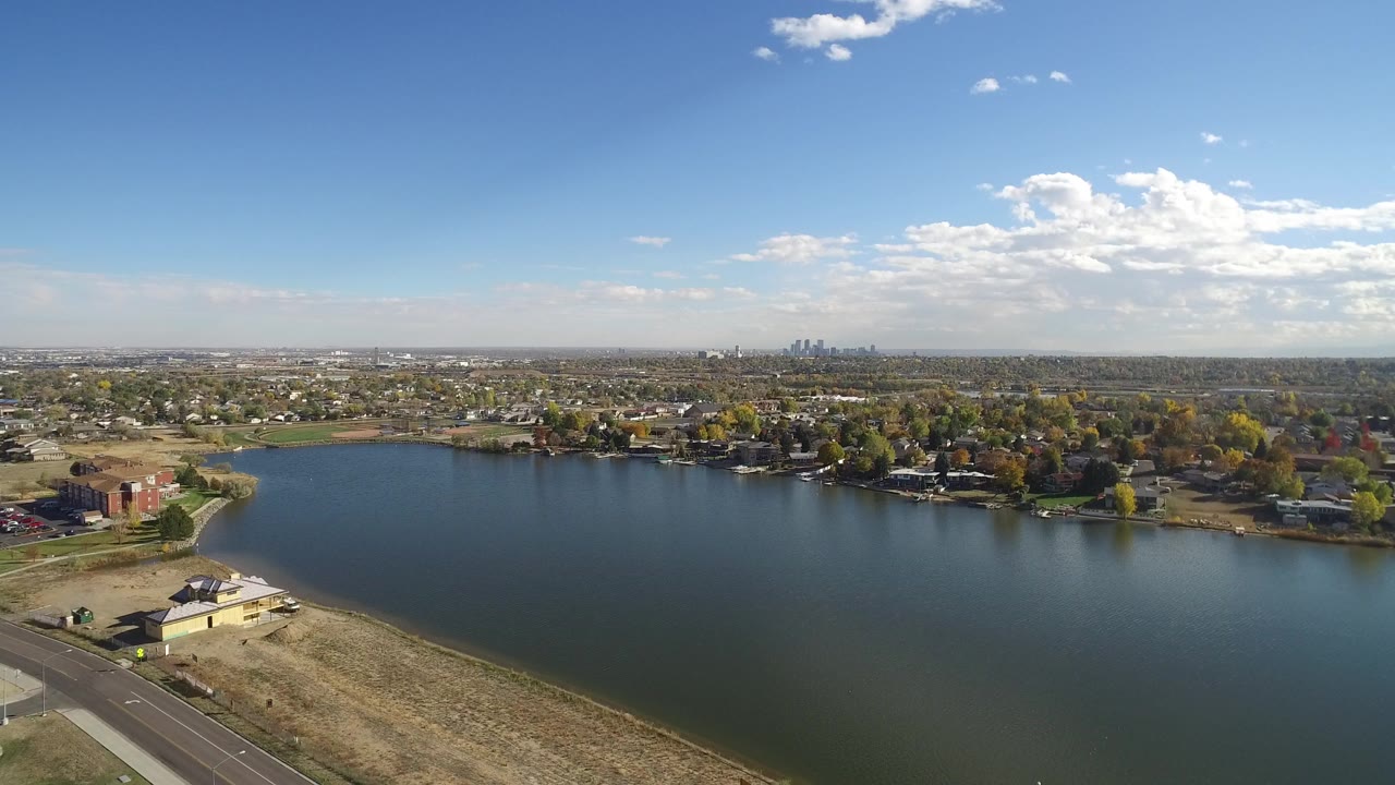 Aerial view over a lake with roads, dry grass, houses, trees, and distant skyline.