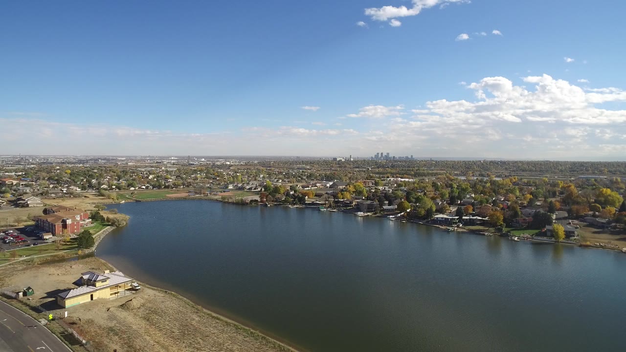 Opening aerial view of Hidden Lake, Colorado, with blue water in the foreground, lakeside homes beyond it, a distant skyline on the horizon, and no visible people.