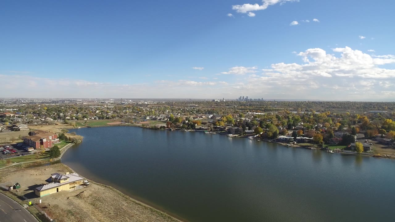Drone-like aerial view across a lake toward homes and a far skyline.