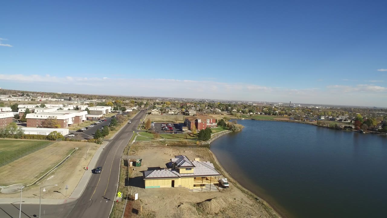 Aerial frame over Hidden Lake, Colorado showing a road at left, a small lakeside construction area in front, blue water to the right, distant low buildings, and no visible people.