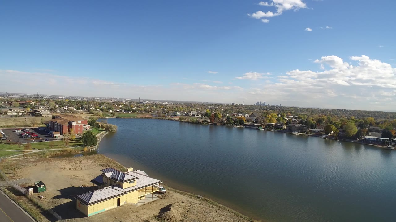 Elevated view over Hidden Lake, Colorado with a sandy shore and small building in the foreground, homes and trees across the water, distant skyline shapes, and no visible people.
